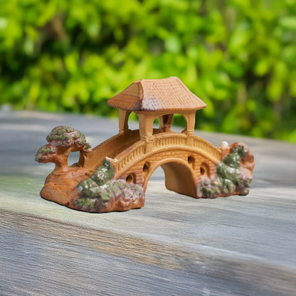 A handcrafted miniature bridge made of brown ceramic, featuring a pagoda-style roof, is displayed on a light wooden surface against a blurred green background.