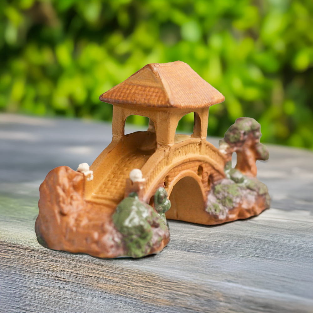 A brown ceramic miniature bridge with a pagoda roof and decorative trees and rocks is set on a wooden surface, viewed from a side angle showcasing its intricate design.