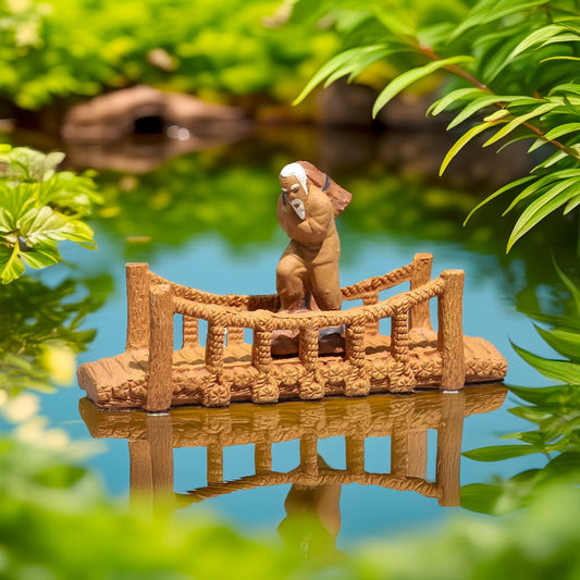 A small ceramic bridge with a rope railing is floating on a pond. A small figure of an old man is standing on the bridge. The water reflects the bridge and the greenery surrounding it.