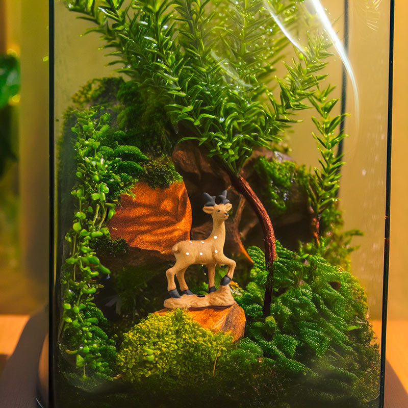 nside a glass terrarium, a miniature deer figurine, hand-painted with exquisite detail, stands amidst a lush green landscape, creating a scene of serene beauty.