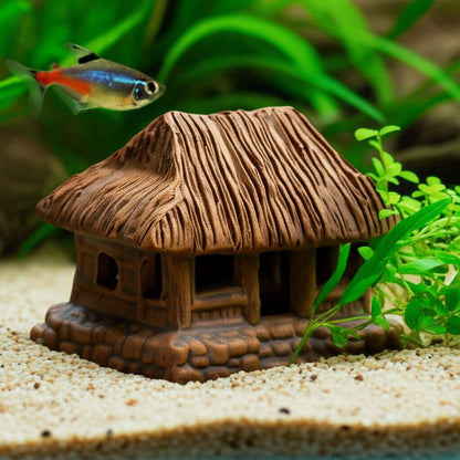 A Thatched House Figurine sits on sand as Ceramic aquarium decor with a small fish nearby.