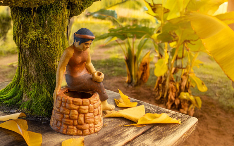 Bathed in golden sunlight, a Vietnamese Tam ceramic figurine sits wistfully atop a well, surrounded by lush foliage and fallen leaves, evoking the magic of ancient folktales.