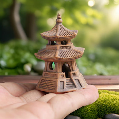 A hand is holding a small brown clay pagoda statue. It has three levels of roofs and open archways on the sides. This is a handcrafted ceramic pagoda figurine.