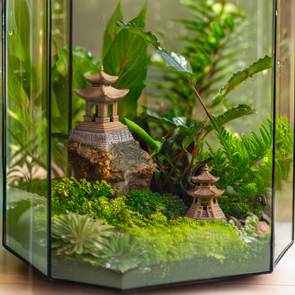 : Two miniature ceramic pagodas, perched atop moss-covered rocks amongst lush ferns in a glass terrarium, transport you to a tranquil Japanese garden.