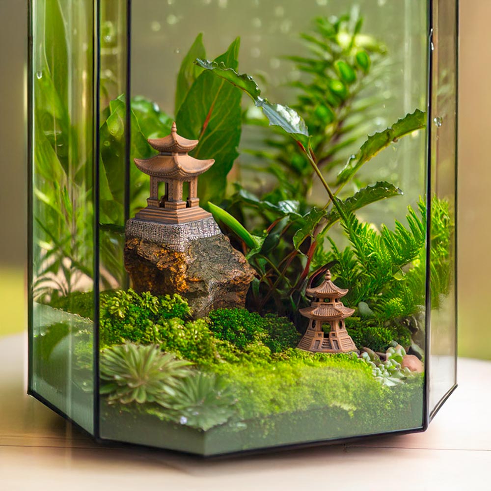  Inside a glass terrarium, two small Bat Trang ceramic pagoda figurines sit among green moss, plants, and a small rock, creating a miniature landscape.