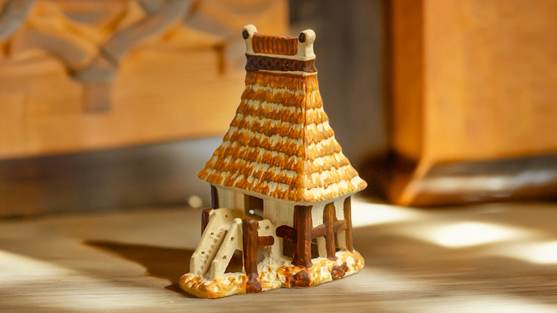 Bathed in warm sunlight, a miniature Vietnamese communal house figurine, meticulously crafted from Bat Trang ceramic, stands proudly on a wooden surface, showcasing the intricate details of its thatched roof and traditional stilt design.