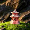 A three-tiered miniature pagoda made of reddish Hoang Tho clay sits atop a lush green moss mound. The ceramic structure features grey-toned roofs and small arched windows, surrounded by smooth black river stones in a zen garden decor setting.