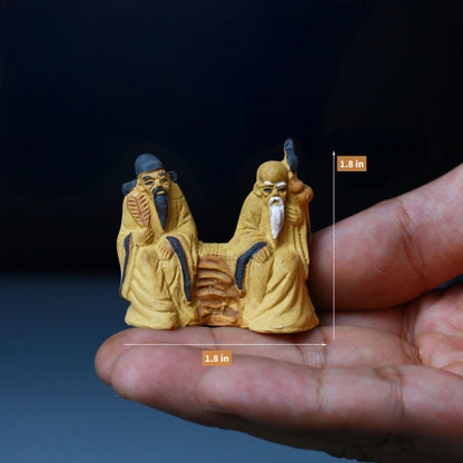 Two small ceramic mudmen figurines are sitting on a bench, held in a hand.