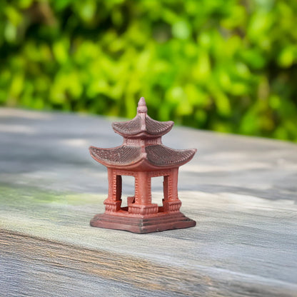 A red ceramic Japanese pagoda with a dark gray roof sits on a gray wooden surface. Its intricate details and vibrant color make it a striking accent for miniature landscapes.