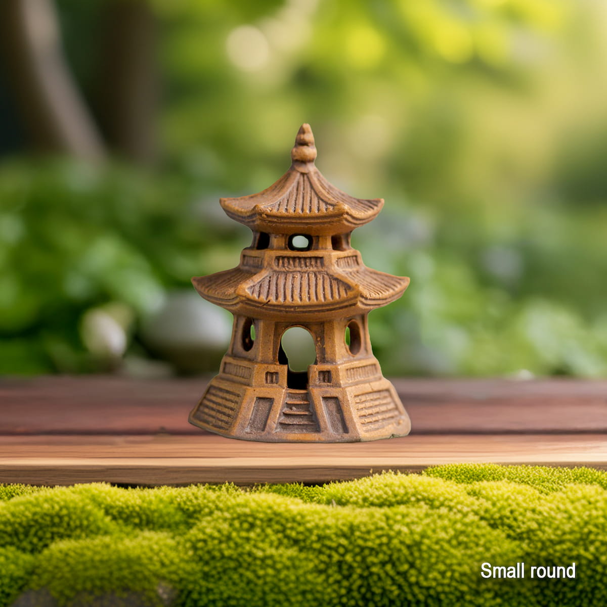 A small round brown clay pagoda statue sits on a wooden surface by green moss. This miniature pagoda figurine is suitable for Zen garden decor.