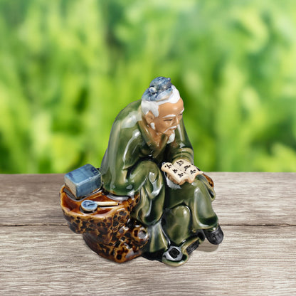 A wise old man, hand-painted in serene shades of green, sits upon a moss-covered rock, lost in the pages of an ancient scroll, adding a touch of wisdom to your bonsai display.