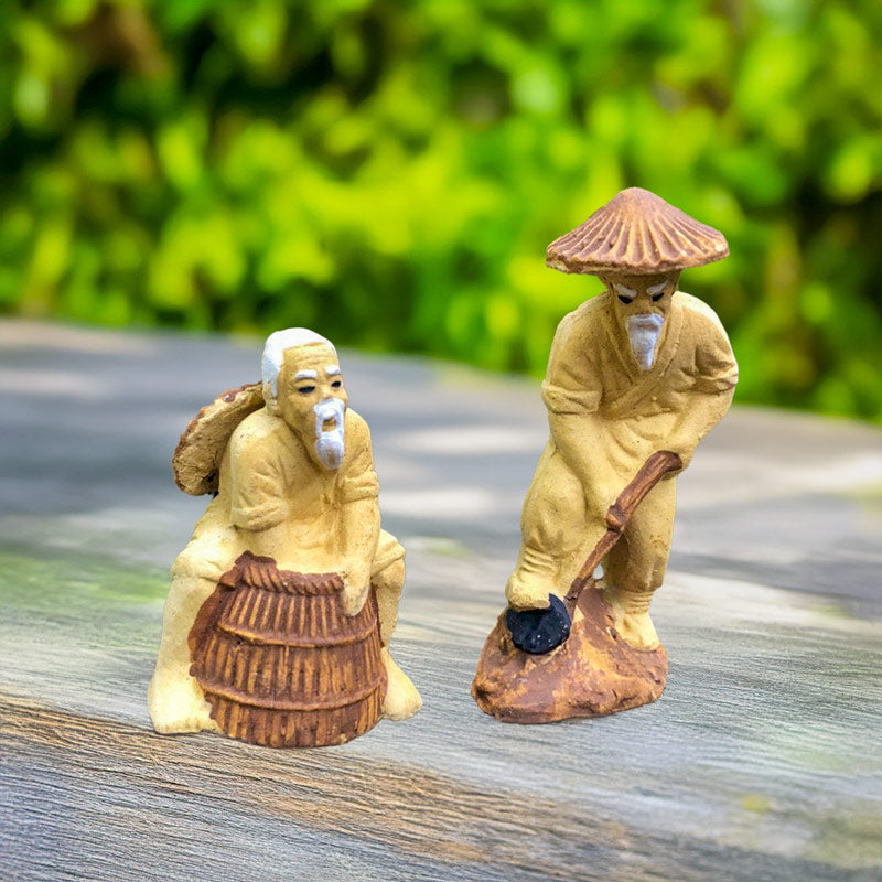 Two Vietnamese Mudmen, handcrafted from natural clay, depict the simple beauty of rural life: one tending his garden, the other fishing with a basket trap.