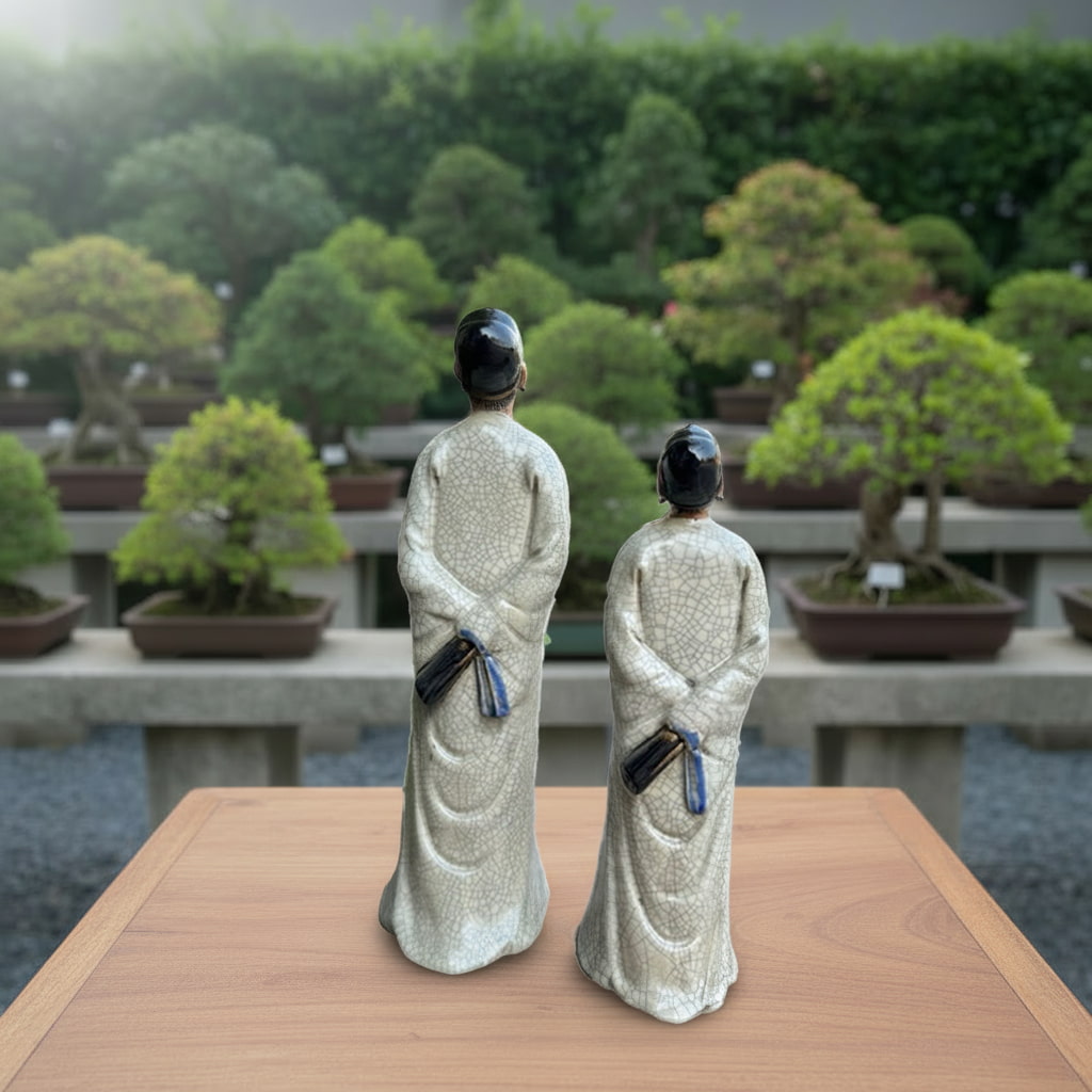 Two miniature bonsai figurines in white crackle-glaze robes, seen from the back on a wooden table.