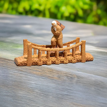 A small ceramic bridge with a rope railing sitting on a wooden surface. There is a small figure of an old man walking across the bridge.