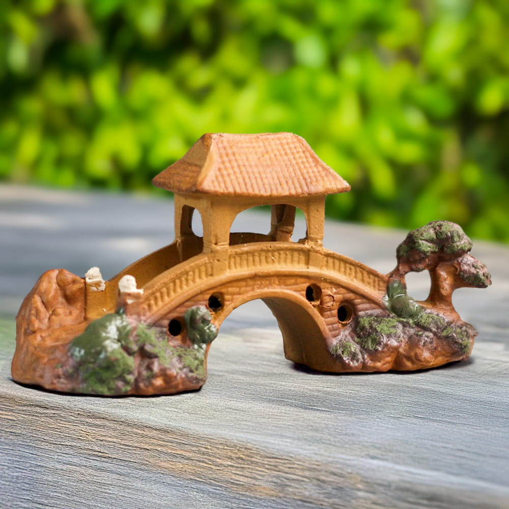 A handcrafted miniature bridge made of brown ceramic, featuring a pagoda-style roof, is displayed on a light wooden surface against a blurred green background.