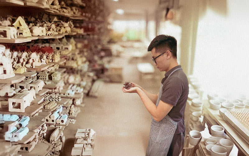 David Nguyen, founder of The Viet Potter, carefully examines a delicate, handmade ceramic figurine in a workshop overflowing with shelves of traditional Bát Tràng ceramic art.