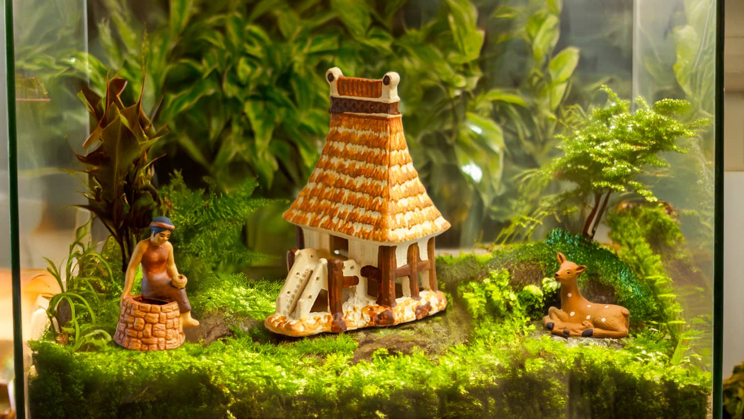 Inside a glass terrarium, a miniature Vietnamese village comes to life. A woman sits by a ceramic well, while a traditional stilt house and a resting deer evoke a sense of tranquility amidst lush greenery. Each piece, handcrafted by artisans in Bat Trang, Vietnam, tells a story of Vietnamese culture and heritage.