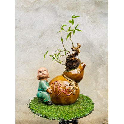 A smiling monk hugs a gourd-shaped figurine bonsai planter. This mudman bonsai pot has a plant.