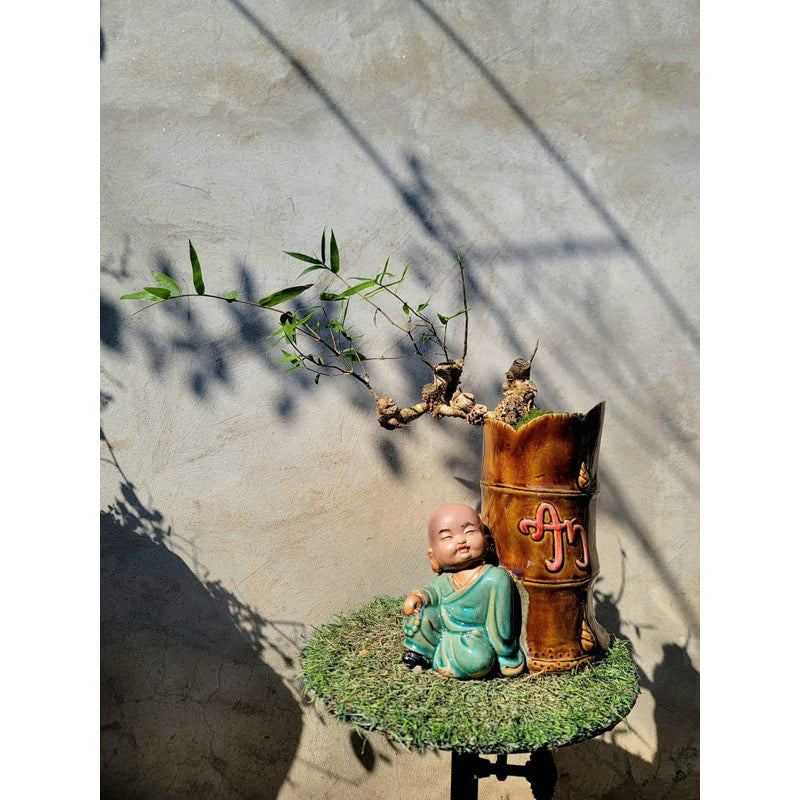  A green-robed mudman leans on a brown, bamboo-shaped mudman bonsai pot with a small plant.