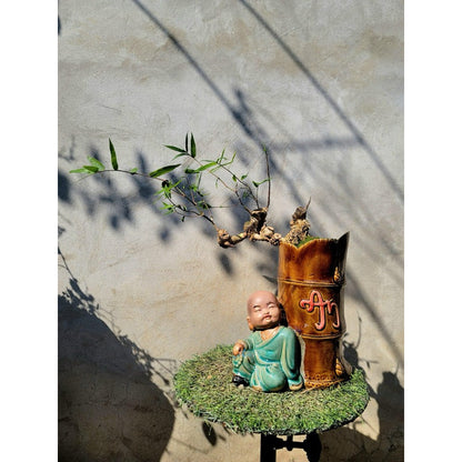  A green-robed mudman leans on a brown, bamboo-shaped mudman bonsai pot with a small plant.