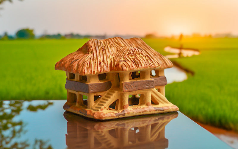 Bathed in the warm glow of sunset, a miniature ceramic Vietnamese stilt house sits on a reflective surface, its intricate details mirrored against a backdrop of verdant rice paddies, evoking the peaceful beauty of Vietnamese countryside.