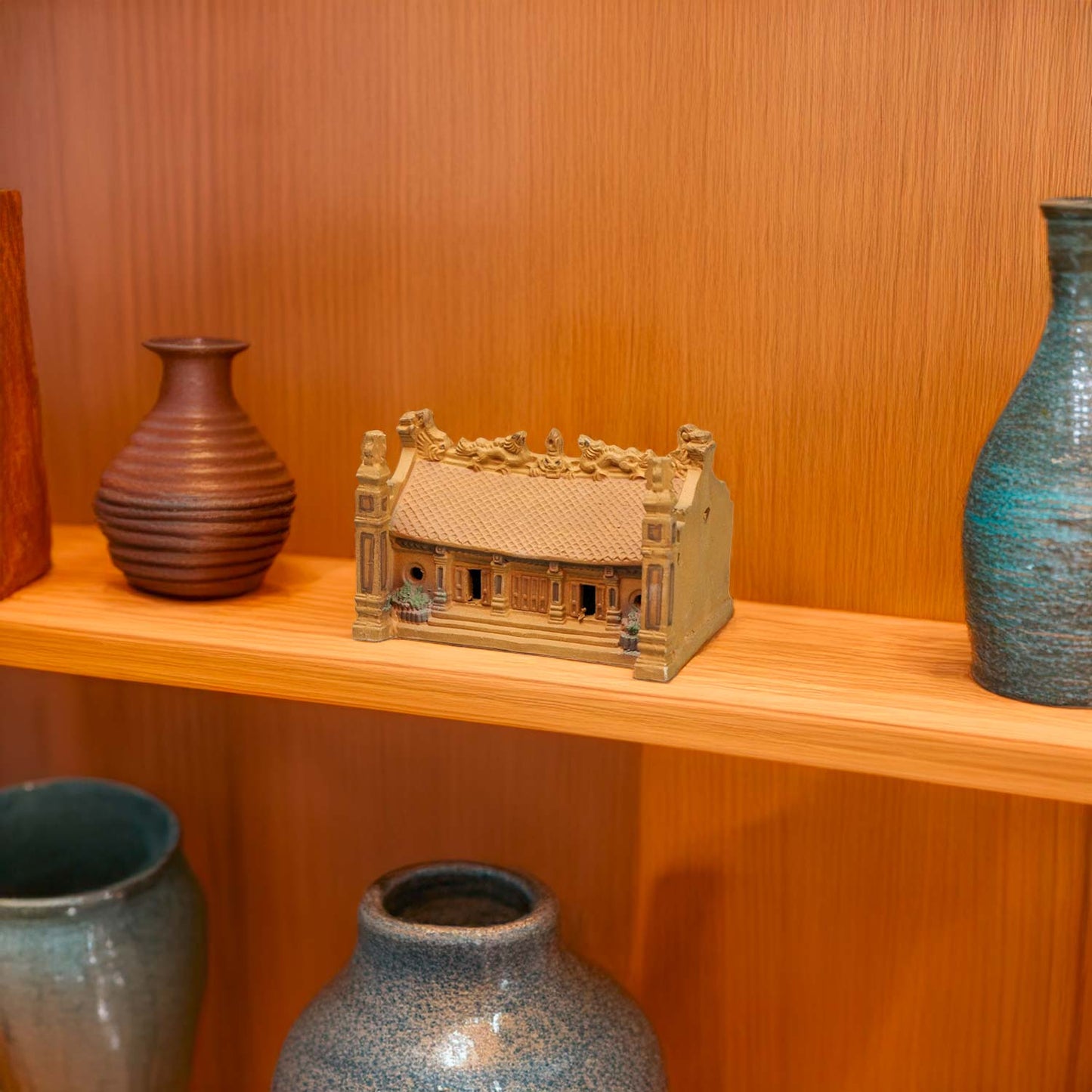 A miniature Vietnamese house, handcrafted from rustic brown ceramic, adds a touch of warmth and cultural charm to a wooden shelf, creating a unique and inviting display.