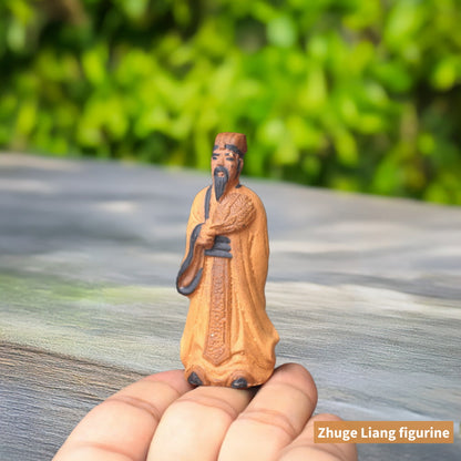 A small, unglazed ceramic figure of Zhuge Liang, a famous Chinese strategist, standing on a person's fingertip. He's wearing a long robe and holding a feather fan.