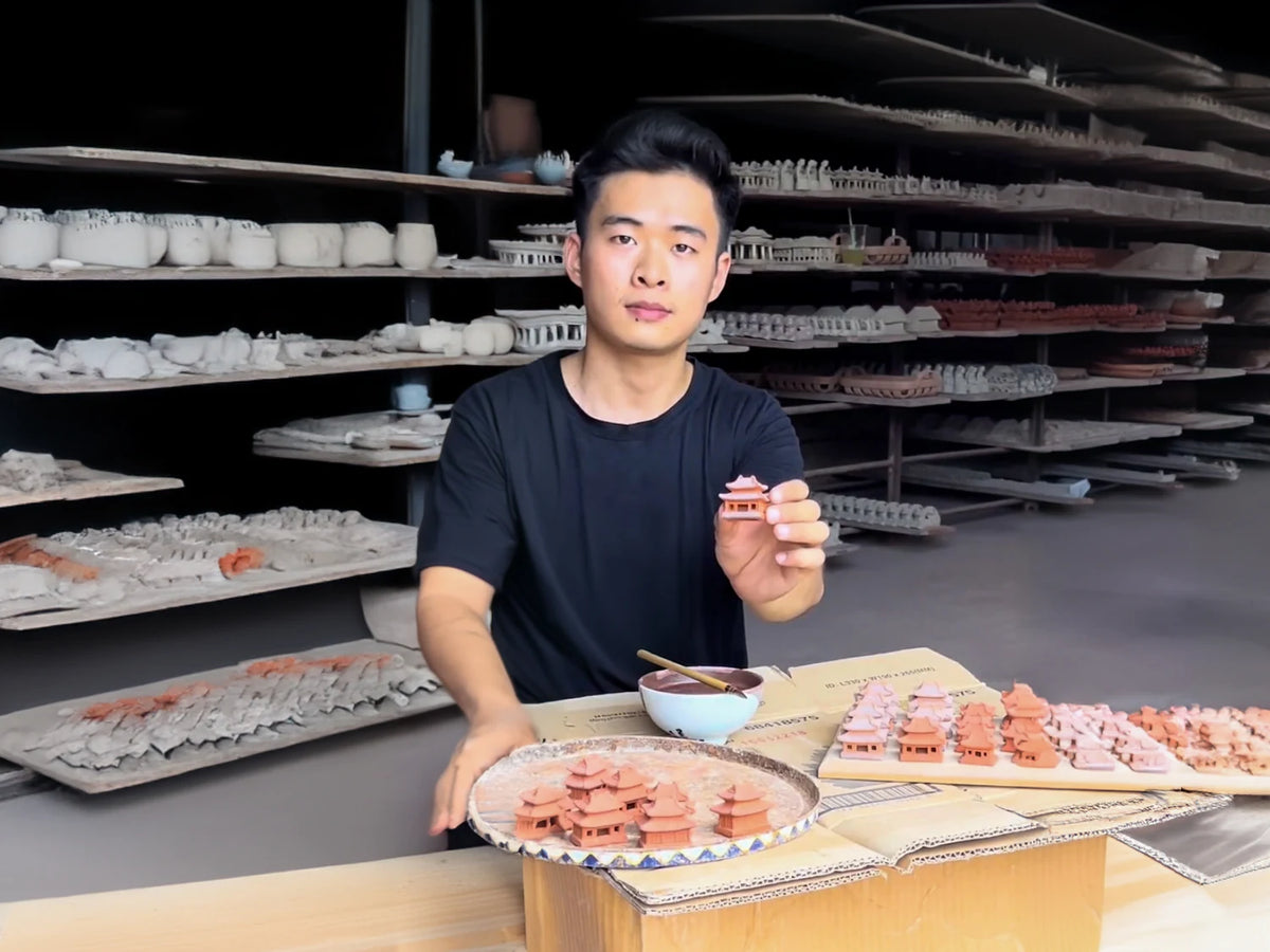 An artisan at Duc Huy Pottery holding a small ceramic figurine in front of shelves of handmade pieces.